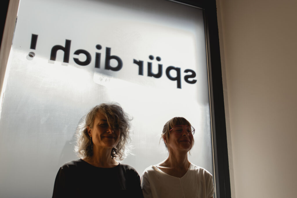 Zwei Frauen sitzen ruhig vor einem Fenster mit dem Schriftzug „spür dich“