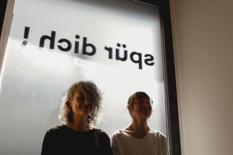 Zwei Frauen sitzen ruhig vor einem Fenster mit dem Schriftzug „spür dich“