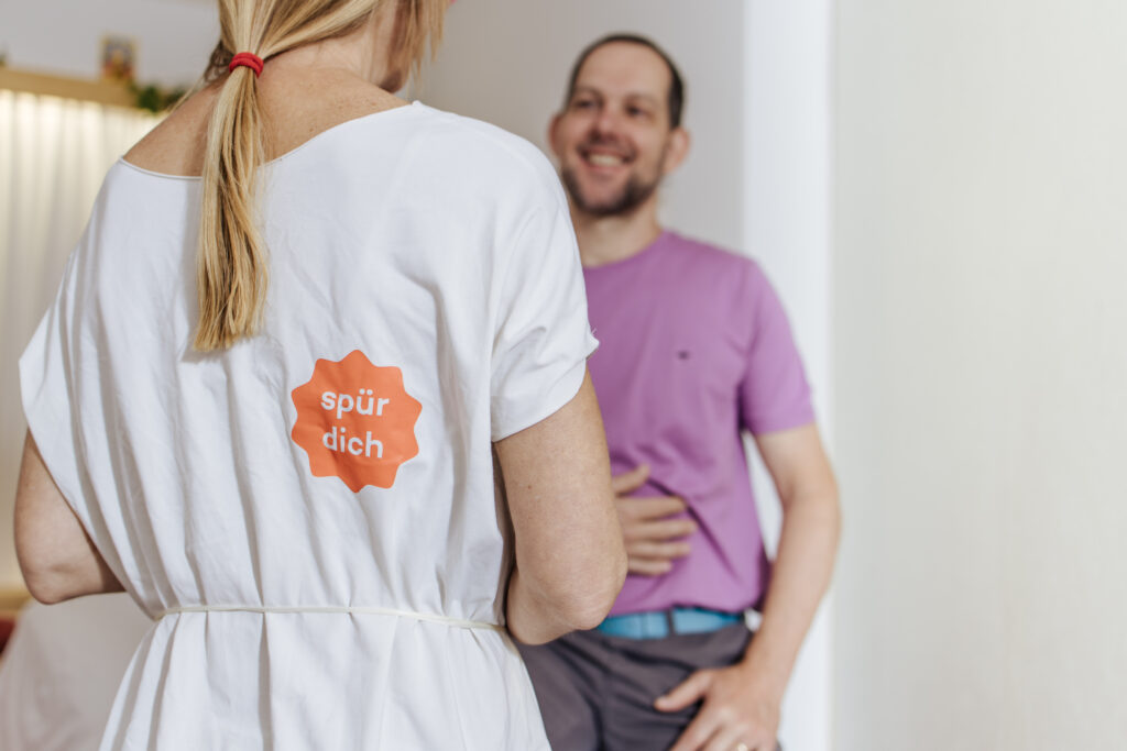 Person mit „spür dich“-Aufdruck im Gespräch mit einem sitzenden Mann
